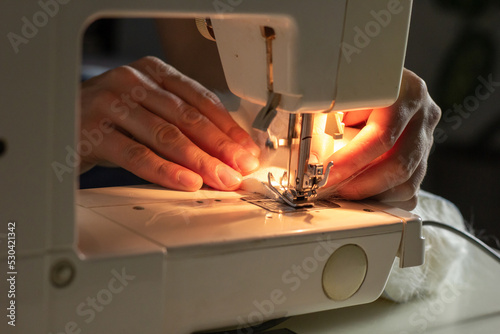 Hands of experienced worker in the handmade industry, sewing white fabric on a sewing machine, the production of Santa red hat. Homemade sewing. Working tailor. Christmas hat. Close up.