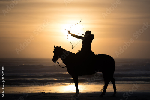 Fotografering Mounted archer holds bow and arrow at sunrise on the beach.