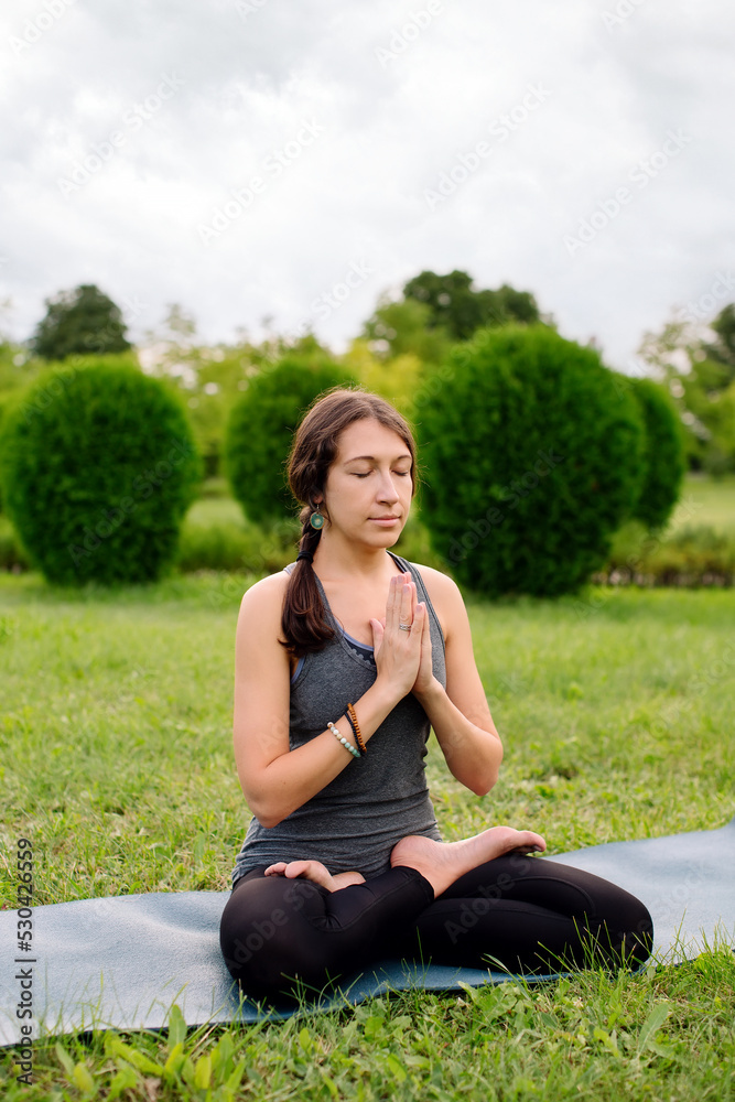 Front view of a woman instructor sitting on yoga mat in lotus position outside in city park. Namaste mudra, seated yoga asana, lotus pose, lotus posture, padmasana, cross legged sitting posture. Front view of a woman instructor sitting on yoga mat in lotus position outside in city park. Namaste mudra, seated yoga asana, lotus pose, lotus posture, padmasana, cross legged sitting posture.