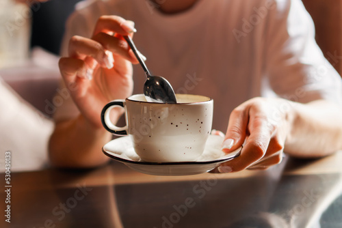 Closeup of female hands with french manicure holding cozy ceramic white mug of tea or coffee. Relax and comfort at home, cafe. Drinking hot cocoa. Empty space for text on blurry background, backplate