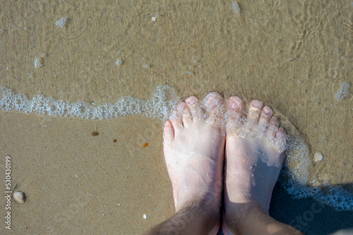 Top view of bare men's feet with approaching seawave on the shore with selective focus, Low angle of of a man standing on the sand beach, Activities and recreation in summer. photo