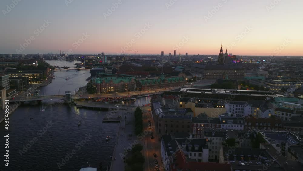 Fly above waterfront at dusk. Buildings and transport infrastructure in evening city. Revealing historic buildings of Borsen and Christiansborg. Copenhagen, Denmark