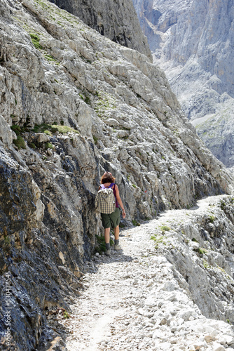 woman hiker with backpack on shoulders walks on stone path in high Dolomites mountain of european alps