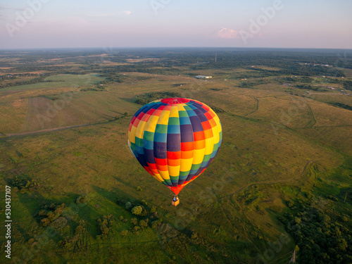 A colorful air balloon is flying in free flight over the field. Bird's-eye view. Multi colored balloon in the blue sky