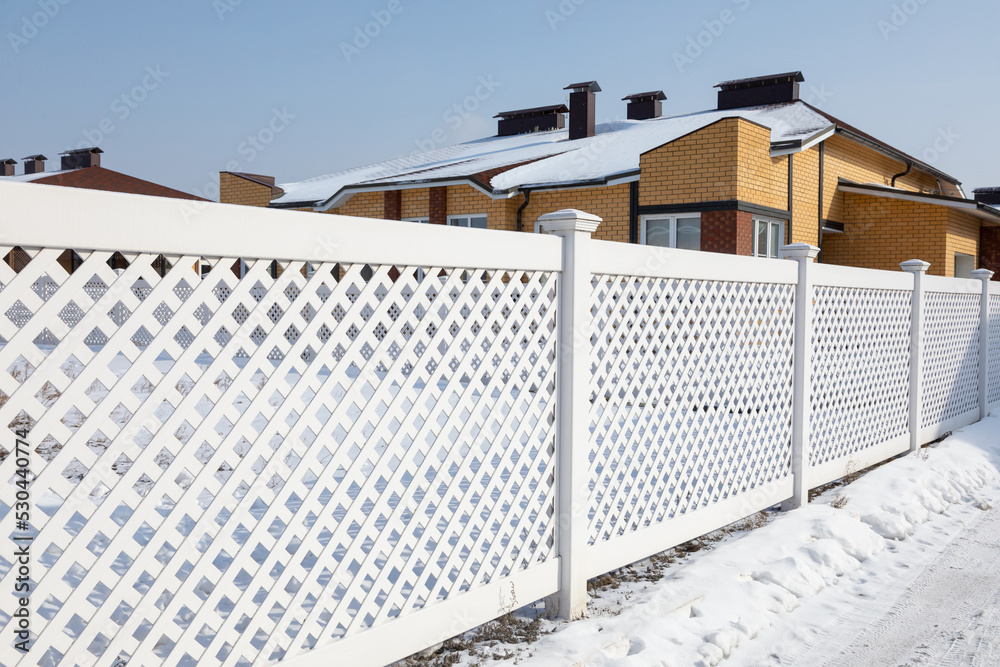 Fototapeta premium White plastic fence in a modern cottage village on a clear winter day. Snow drifts in front of a vinyl fence against a blue sky, a roof of a cottage is covered with snow