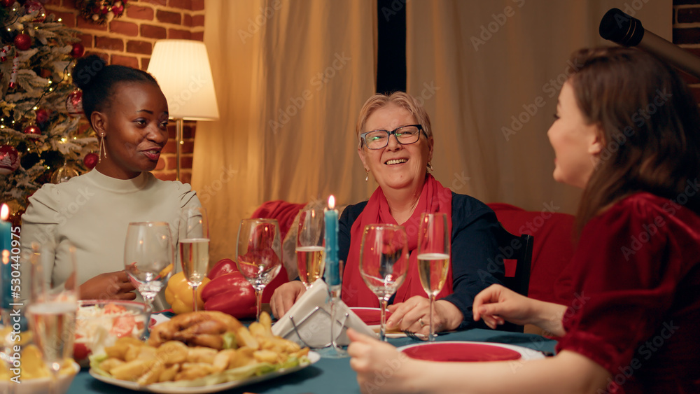 Happy festive women sitting at Christmas dinner table while laughing and talking together. Joyful people gathered in living room at home to celebrate winder season traditional holiday.
