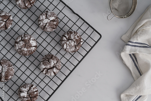 Top down shot food photo of freshly baked Chocolate Crinkle Cookies. Chocolate iced biscuits arranged on black wire cooling rack. White work surface background. Copy space available
