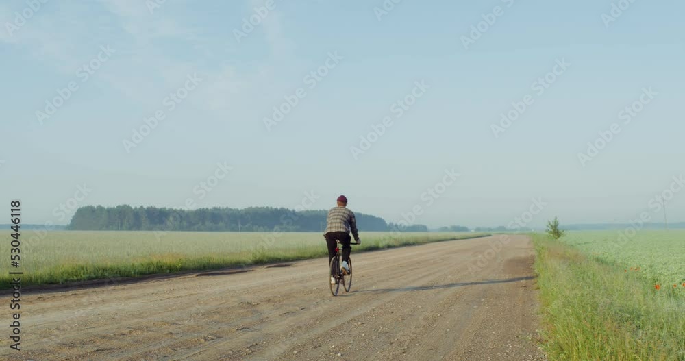 A young, stylishly dressed man rides a bicycle on a gravel road near a wheat field, illuminated by bright sunlight. Video from the back. 4k video, red komodo A young, stylishly dressed man rides a bicycle on a gravel road near a wheat field, illuminated by bright sunlight. Video from the back. 4k video, red komodo