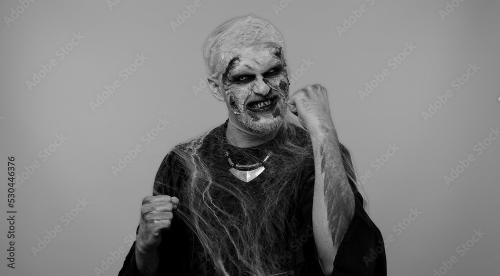 Spooky crazy zombie man alone with makeup with fake wounds scars celebrate success win scream rejoices doing winner hands gesture say Yes in gray studio. Sinister dead guy. Halloween holidays Spooky crazy zombie man alone with makeup with fake wounds scars celebrate success win scream rejoices doing winner hands gesture say Yes in gray studio. Sinister dead guy. Halloween holidays