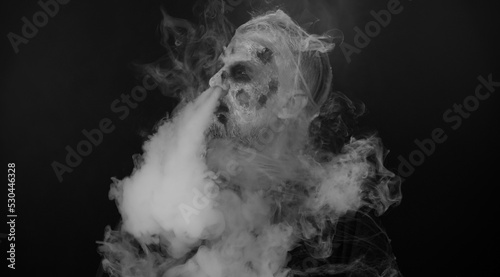 Frightening man with Halloween zombie bloody wounded make-up hiding behind hands, blows smoke from nose and mouth. Horror theme. Sinister undead guy isolated on studio black background. Voodoo rituals