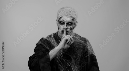 Shh be quiet. Sinister man with horrible scary Halloween zombie makeup in costume presses index finger to lips makes silence gesture sign do not tells secret. Dead guy with wounded bloody scars face