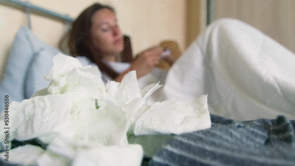 Sick woman lies in bed, takes her temperature and drinks hot tea in soft focus, pile of used handkerchiefs in the foreground. Suffering from seasonal flu or cold Sick woman lies in bed, takes her temperature and drinks hot tea in soft focus, pile of used handkerchiefs in the foreground. Suffering from seasonal flu or cold