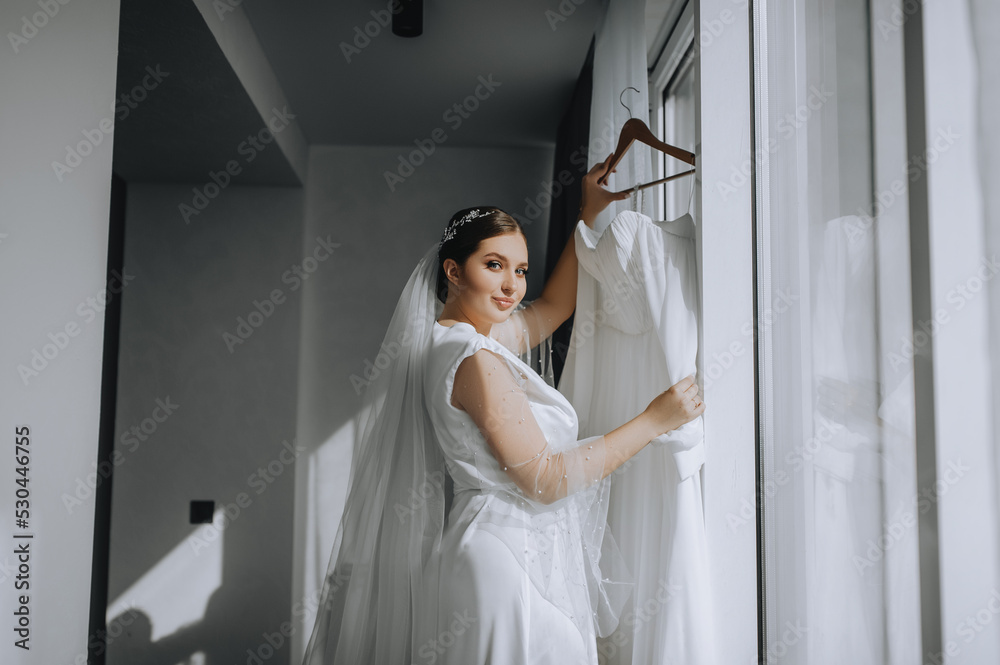 A beautiful brunette bride model in lingerie, a white bathrobe stands near the window in the morning in a hotel, indoors and holds a dress on a hanger. Wedding portrait, photography. A beautiful brunette bride model in lingerie, a white bathrobe stands near the window in the morning in a hotel, indoors and holds a dress on a hanger. Wedding portrait, photography.