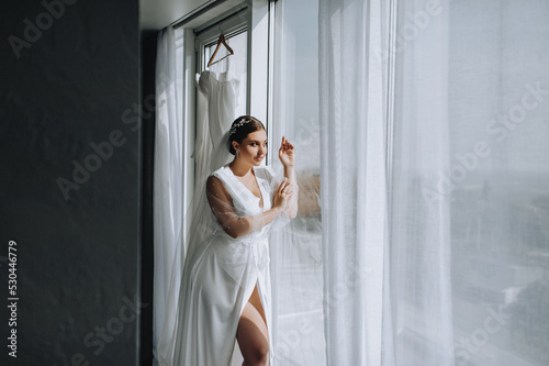 A beautiful brunette bride model in lingerie, a white bathrobe stands near the window in the morning. Wedding portrait, photography.