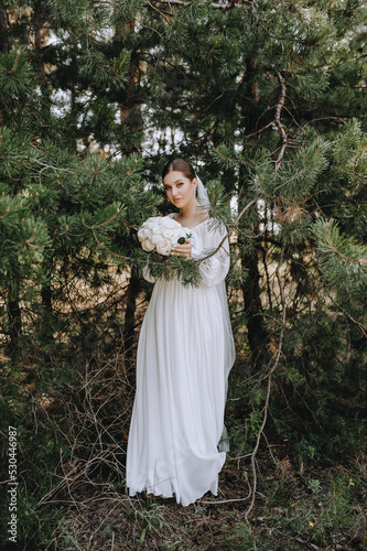 A beautiful, young brunette bride in a white long dress with a bouquet of roses stands in a coniferous forest in nature near a green spruce. Wedding portrait, photography.