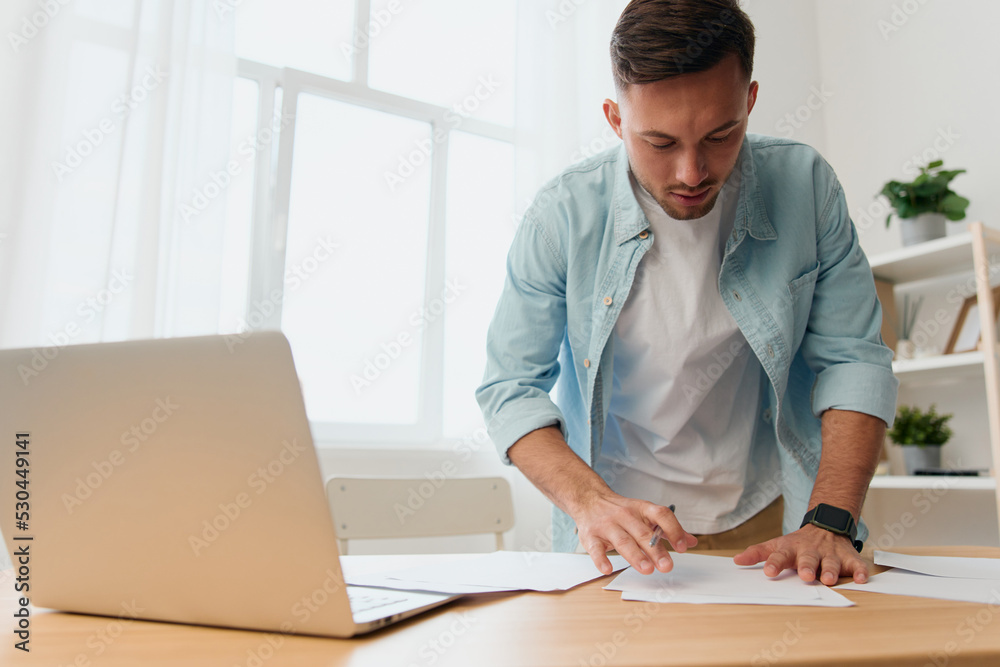 Thoughtful pensive handsome stylish young businessman deals with documents and papers for work project on office table. Copy space for ad. Remote Job, Technology And Career Profession Concept Thoughtful pensive handsome stylish young businessman deals with documents and papers for work project on office table. Copy space for ad. Remote Job, Technology And Career Profession Concept