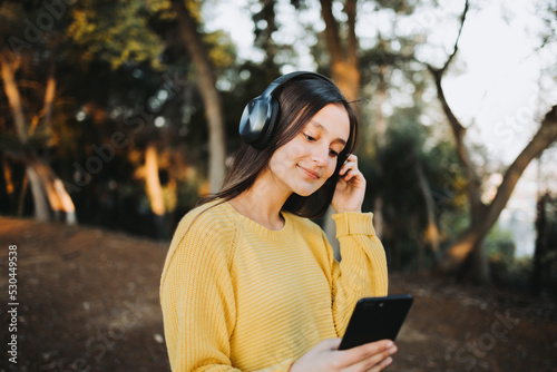 Teenage girl wearing yellow sweater, using headphones for playing music on her smartphone in the park photo
