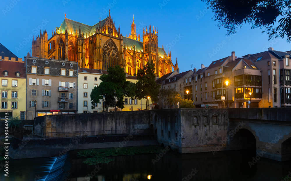 Obraz premium Scenic nighttime view of summer Metz cityscape overlooking illuminated majestic Gothic Roman Catholic Cathedral towering over residential buildings on waterfront along Moselle River, France
