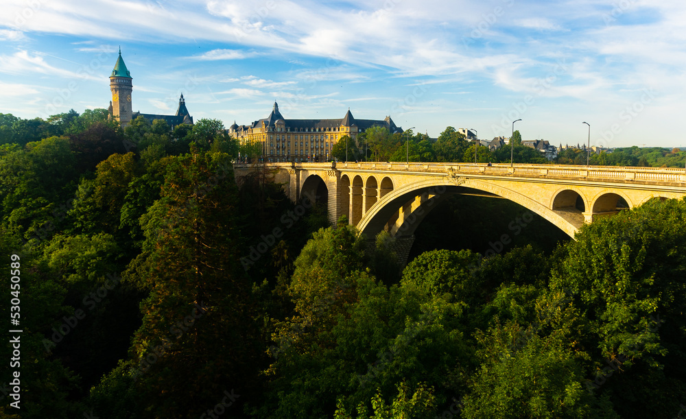 Scenic view of Adolphe Bridge, stone arched aqueduct running over green valley of Petrusse River in Luxembourg city with State Savings Bank and banking museum in background on sunny summer day..