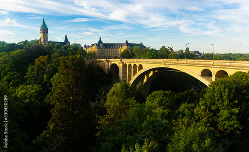 Scenic view of Adolphe Bridge, stone arched aqueduct running over green valley of Petrusse River in Luxembourg city with State Savings Bank and banking museum in background on sunny summer day.. photo