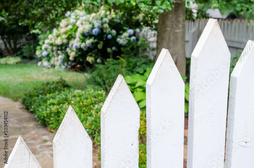 A white picket fence with flower garden in the distance
