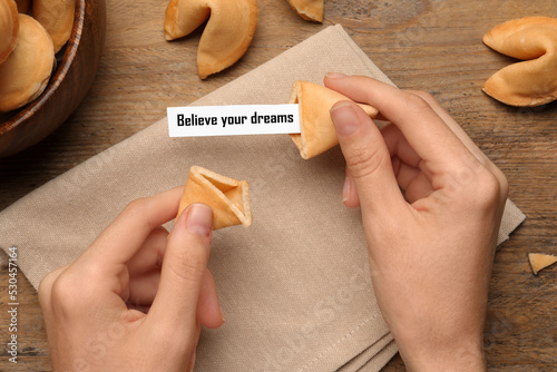 Woman holding tasty fortune cookie with prediction Believe your dreams at wooden table, top view photo