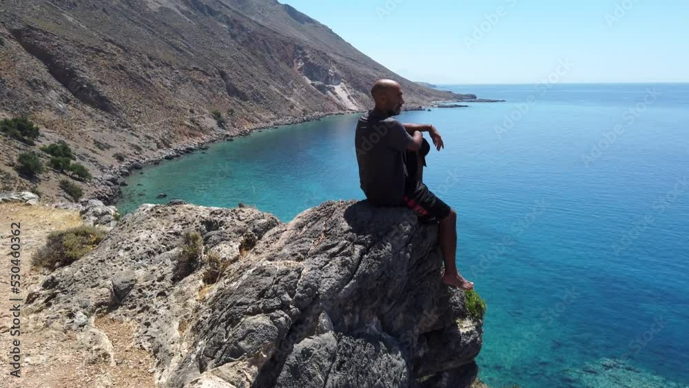 Barefoot meditating man sitting on the edge of a cliff contemplating the panoramic view of the amphitheater bay of Loutro