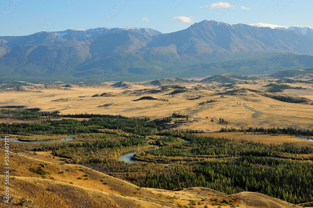 The winding bed of a beautiful river flowing through the hilly steppe at the foot of high mountains in early autumn. The winding bed of a beautiful river flowing through the hilly steppe at the foot of high mountains in early autumn.