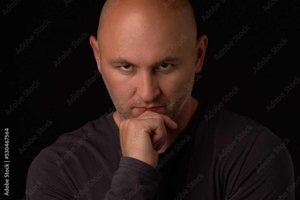 Bald, unshaven man in a black jacket looks thoughtfully into the camera and holds his hand on his chin, a photo on a black background