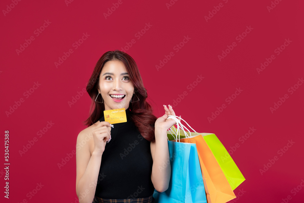Obraz premium Stylish Asian woman in black dress holding shopping bags holding gold credit cards on crimson background. Beautiful Thai woman holding shopping bags holding gold credit cards on dark red background.