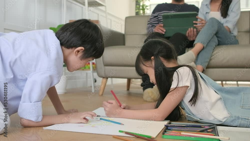 Asian Thai siblings are lying on living room floor, drawing homework with colored pencils together, parents leisurely relax on a sofa, lovely happy weekend activity, and domestic wellbeing lifestyle. Asian Thai siblings are lying on living room floor, drawing homework with colored pencils together, parents leisurely relax on a sofa, lovely happy weekend activity, and domestic wellbeing lifestyle.