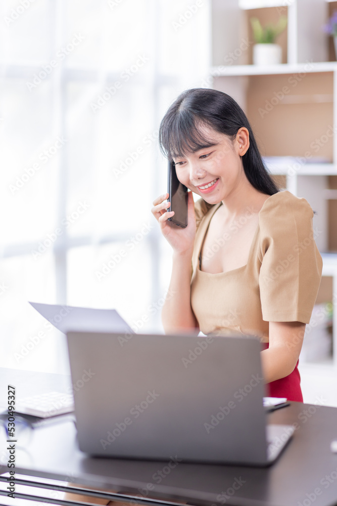 Portrait of young Asian smiling cheerful entrepreneur in casual office making a phone call while working with a laptop in her workplace Portrait of young Asian smiling cheerful entrepreneur in casual office making a phone call while working with a laptop in her workplace