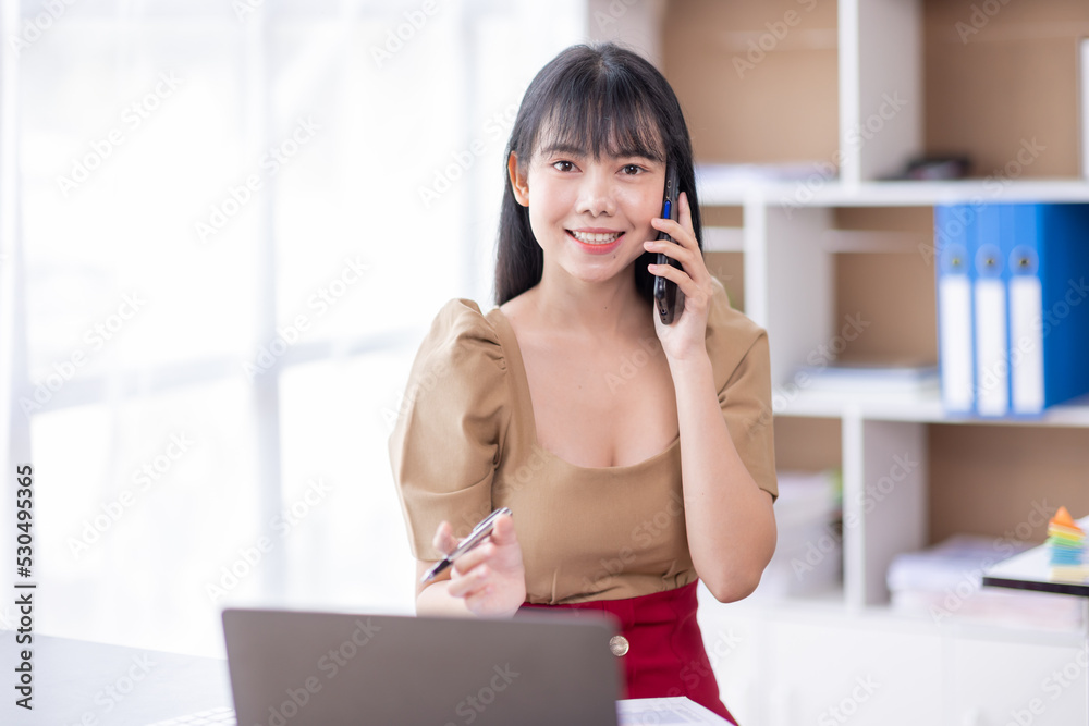 Portrait of young Asian smiling cheerful entrepreneur in casual office making a phone call while working with a laptop in her workplace Portrait of young Asian smiling cheerful entrepreneur in casual office making a phone call while working with a laptop in her workplace