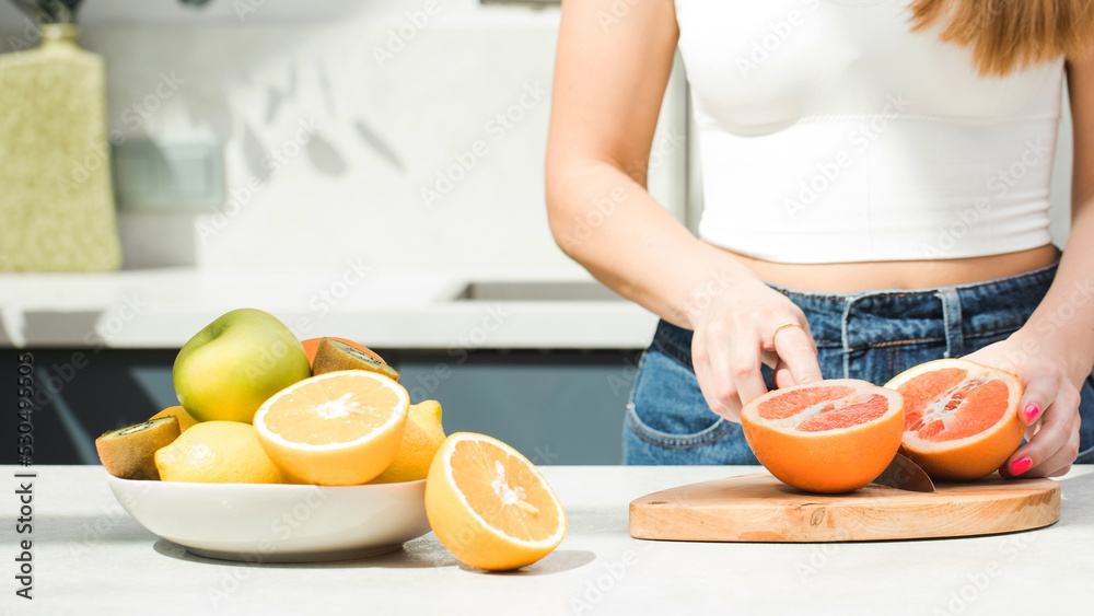 A woman cuts a grapefruit at the table in the kitchen, a plate with fresh fruits. Healthy food concept. A woman cuts a grapefruit at the table in the kitchen, a plate with fresh fruits. Healthy food concept.