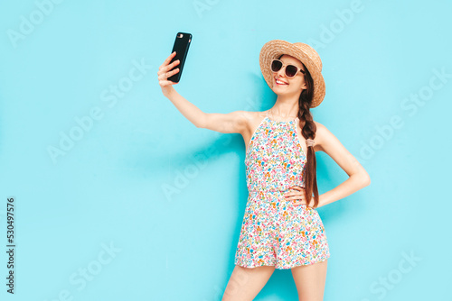 Portrait of young beautiful smiling female in trendy summer overalls. carefree woman posing near blue wall in studio. Positive model having fun indoors. Cheerful and happy. In hat. Taking selfie