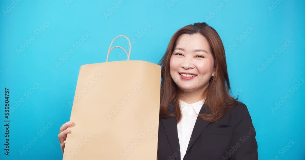 Beautiful Asian smiling businesswoman in a black suit with a shopping bag holding and cheerful blue background Beautiful Asian smiling businesswoman in a black suit with a shopping bag holding and cheerful blue background