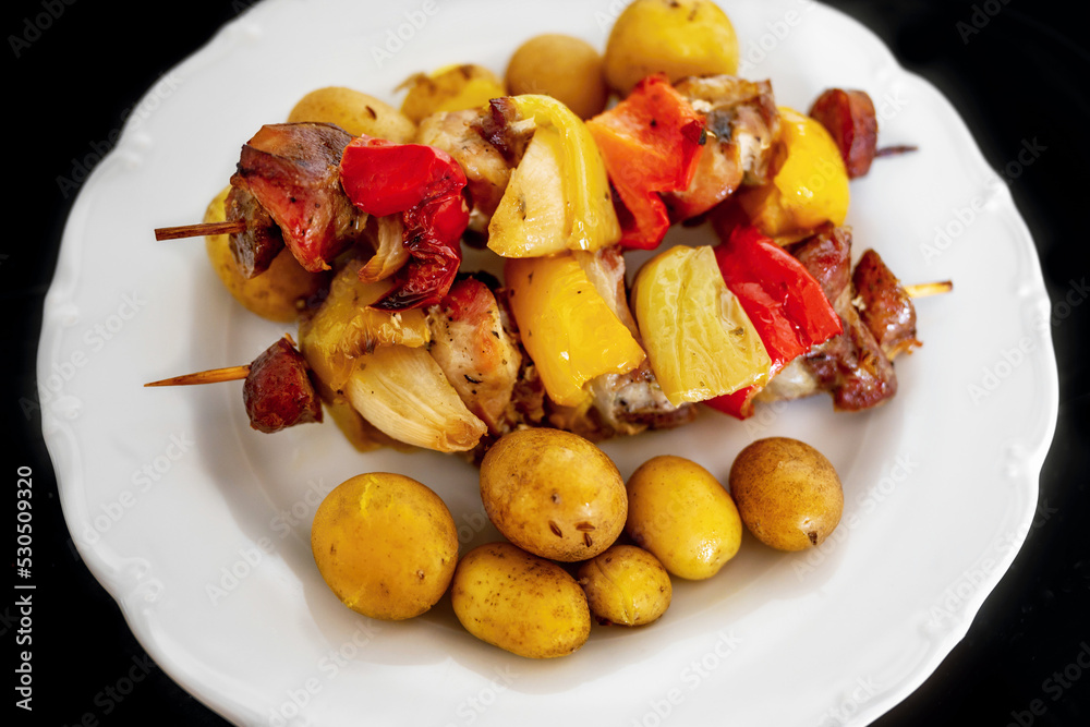 Skewer with pieces of pork meat,paprika,sausage,bacon and onion and boiled potato on plate.