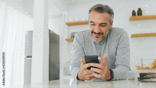 Attractive Middle-aged man using mobile phone or smartphone while standing in kitchen at home.