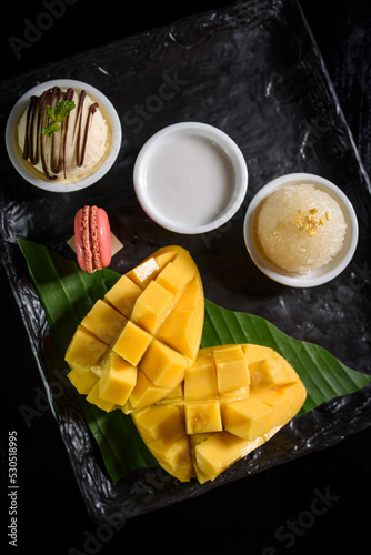 Fototapeta Naklejka Na Ścianę i Meble -  Mango sticky rice, coconut milk, ice cream and macarons on a black plate Popular Thai desserts in summer