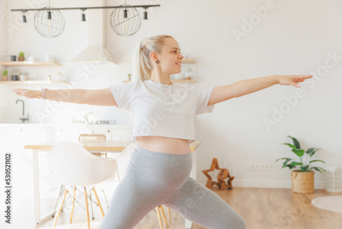 Concept fitness pregnancy. Happy scandy pregnant blond woman doing sports stretching on mat background white kitchen