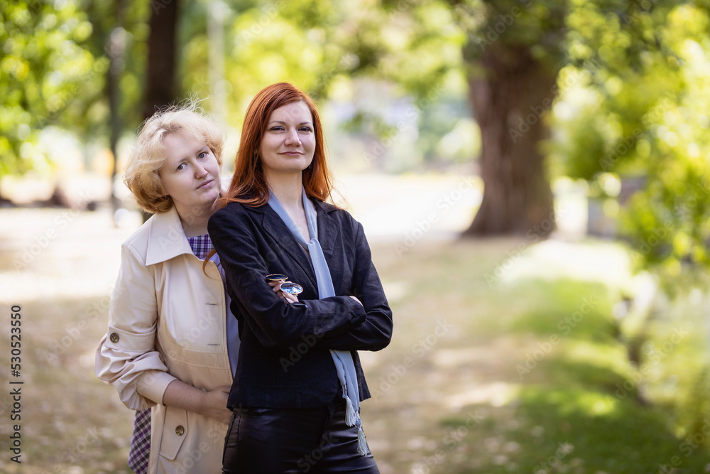 Fototapeta premium Two women together - blonde in light and redhead in dark clothes posing in a park. Cosplay - female version of Angel and Demon (Good Omens)