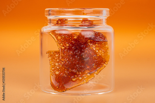 glass jar with  brown cannabis wax, pieces of hardened resin of marijuana extract on an orange background.