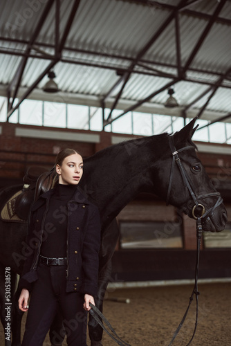 Woman jockey in a black uniform with his black horse inside the dark indoor manege. Arena horse. Equestrian sport. Racecourse. Sport and fashion concept. © Oksana