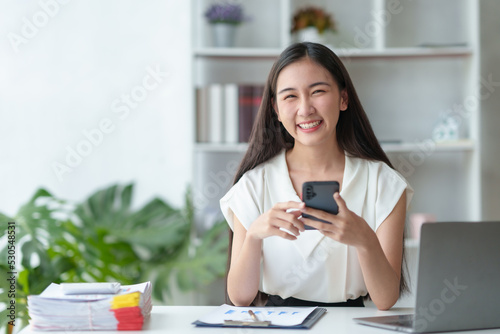 Beautiful Asian businesswoman using a smartphone to contact customers reply to chat Transact on social media applications in the office with a smiling face.