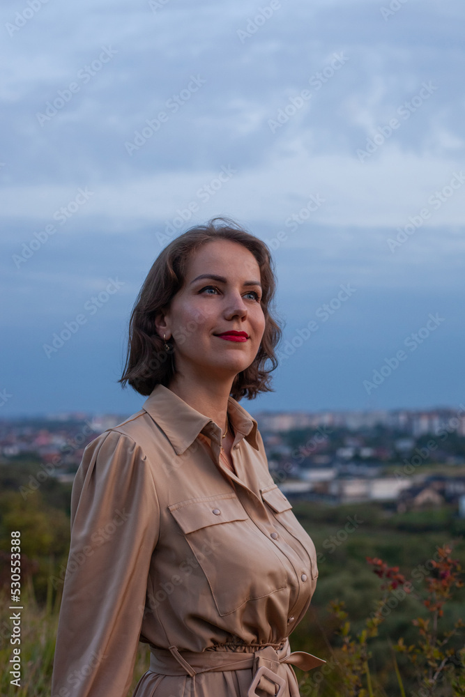 Portrait of beautiful young smiling girl in a long brown dress with sky background. Happy woman stands at sunset in a hill garden. Concept of having rest in park during summer holidays or weekends