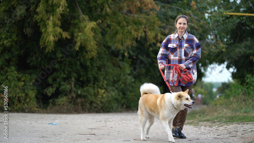 Woman in plaid shirt holding akita inu dog on red leash while walking outdoors