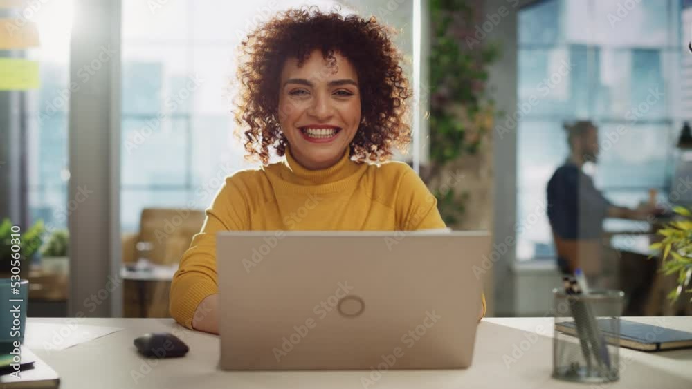 Portrait of a Beautiful Middle Eastern Manager Sitting at a Desk in Creative Office. Young Stylish Female with Curly Hair Looking at Camera with Big Smile. Colleagues Working in the Background.