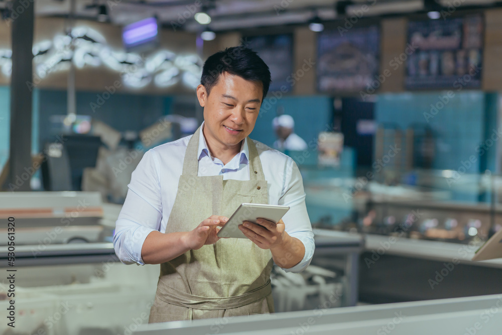 Asian grocery store manager salesman in apron using digital tablet counting goods in supermarket worker employee doing inspection of products regular inventory to control goods online order Asian grocery store manager salesman in apron using digital tablet counting goods in supermarket worker employee doing inspection of products regular inventory to control goods online order