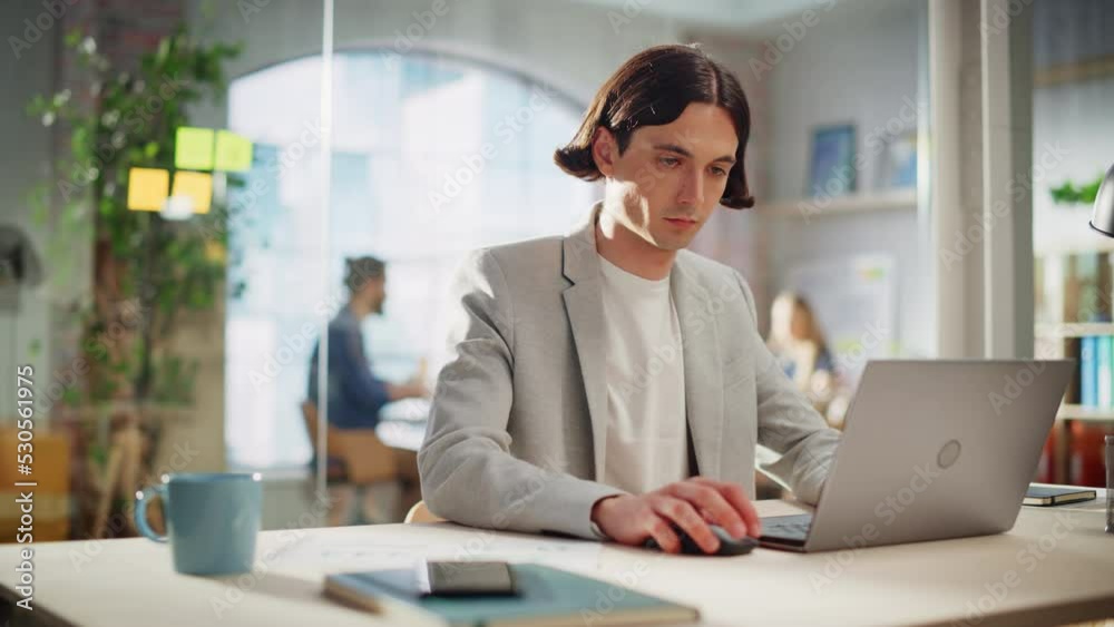 Handsome Manager Sitting at a Desk in Creative Office. Young Stylish Man in a Jacket Using Laptop Computer in Marketing Agency. Colleagues Working in the Background. Handsome Manager Sitting at a Desk in Creative Office. Young Stylish Man in a Jacket Using Laptop Computer in Marketing Agency. Colleagues Working in the Background.