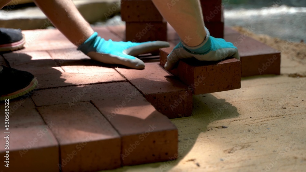 Low angle extreme closeup of person installing brick pavers onto sand base wearing blue gloves. Low angle extreme closeup of person installing brick pavers onto sand base wearing blue gloves.